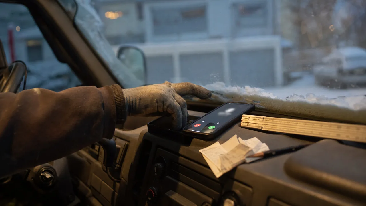 Hånd i arbeidshanske strekker seg etter en mobil som ringer på dashbordet i en varebil, med vintermørke og snø utenfor frontruta
