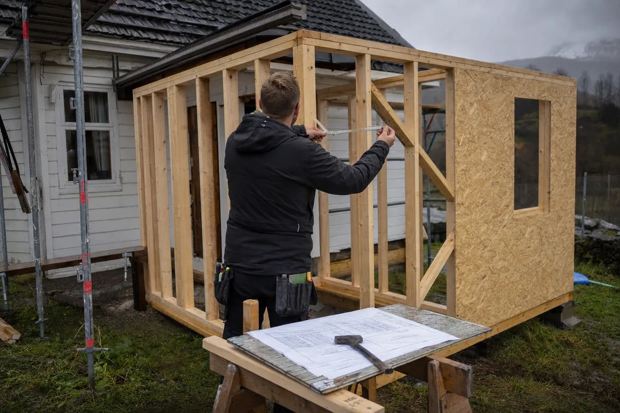 Bindingsverk til et tilbygg på et norsk trehus, med en tømrer som måler og tegninger lagt utover en bukk